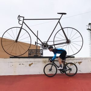 Road cyclist riding on the red track of the Sineu velodrome beneath a large metal bicycle sculpture