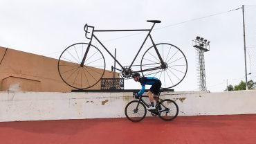 Road cyclist riding on the red track of the Sineu velodrome beneath a large metal bicycle sculpture